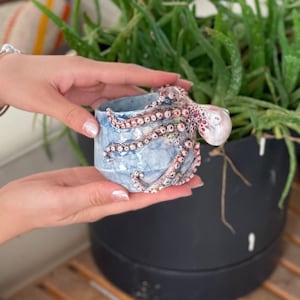 Könnte beinhalten: Ein kleiner blauer Keramiktopf mit einem rosa-weißen Kraken-Design. Der Topf wird von einer Hand gehalten.