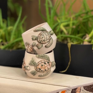 May include: Two white ceramic mugs featuring a 3D design of sea turtles. The turtles are green, brown, and beige. The mugs are stacked on top of each other.