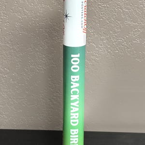 May include: A cylindrical cardboard shipping tube with a white top section featuring the red text Scratchoff Posters.com and a black star icon. The lower section is green with the white text 100 BACKYARD BIRDS printed vertically along the side.