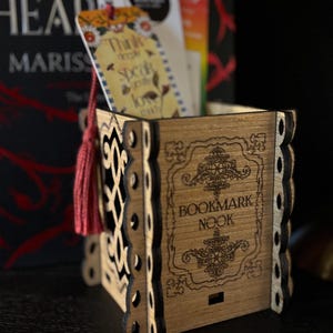 Op de afbeelding: Een houten boekenleggerhouder met de woorden "Bookmark Nook" gegraveerd op de voorkant. De houder heeft een open ontwerp aan de zijkanten en een rode kwast. Binnenin zitten verschillende bladwijzers, één met de tekst "Think deeply, speak gently, love much."