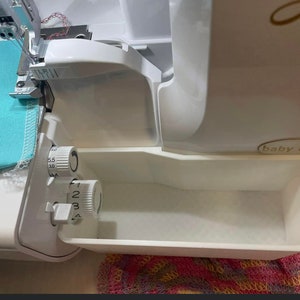 Op de afbeelding: Een witte Baby Lock lockmachine met lichtblauwe stof die wordt genaaid. De machine heeft verstelbare draaiknoppen en een ingebouwde afvalbak. Ontworpen voor het naaien en afwerken van stofkanten.