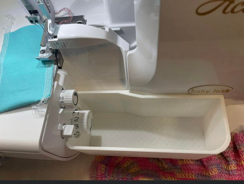 Op de afbeelding: Een witte Baby Lock lockmachine met lichtblauwe stof die wordt genaaid. De machine heeft verstelbare draaiknoppen en een ingebouwde afvalbak. Ontworpen voor het naaien en afwerken van stofkanten.