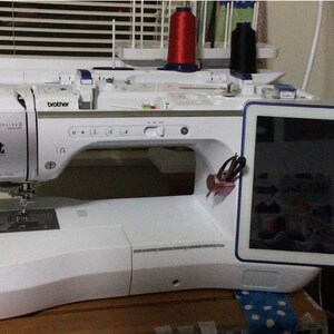 May include: A white Brother Luminaire 2 sewing and embroidery machine featuring a large digital touchscreen display on the right side, a needle area, and a top-mounted thread spool holder with red and black thread spools positioned above the machine.
