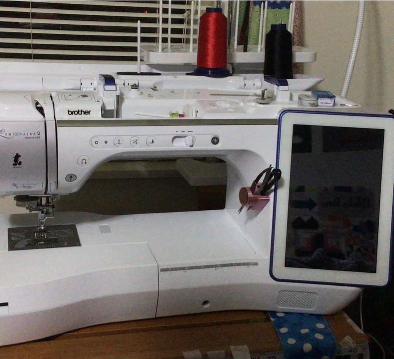 May include: A white Brother Luminaire 2 sewing and embroidery machine featuring a large digital touchscreen display on the right side, a needle area, and a top-mounted thread spool holder with red and black thread spools positioned above the machine.