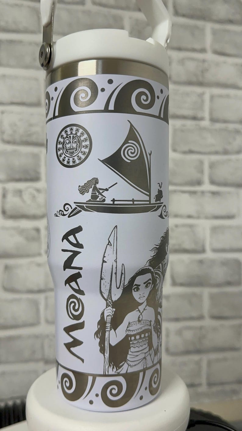 May include: White stainless steel travel mug with a black and white design featuring Moana from the Disney movie Moana. The design includes Moana, Maui, and the ocean.