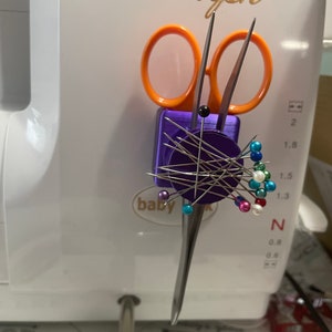 May include: A purple magnetic pincushion with a variety of coloured pins and two pairs of scissors. The pincushion is attached to a white sewing machine with the word "Triumph" written in gold lettering.
