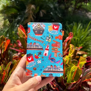 May include: A blue travel wallet with a pattern of cruise ships, Mickey Mouse, Stitch, and other nautical elements. The wallet has the words "Castaway Cay" printed on it. The background is a lush green and red foliage.