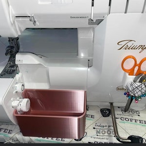 Op de afbeelding: Een witte Baby Lock Triumph lockmachine met ros&eacute;gouden opbergbakken. De machine heeft een oranje schaar en spelden in een houder. De tekst "Triumph" staat in goud op de machine.