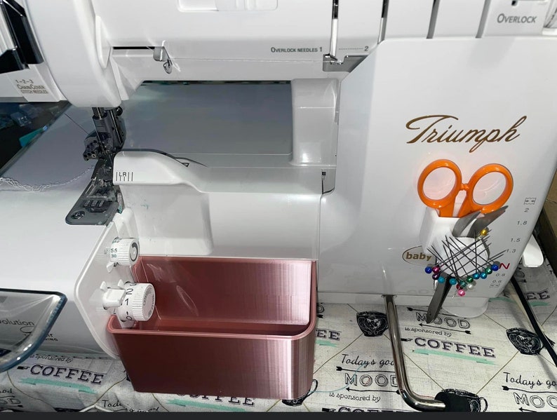Op de afbeelding: Een witte Baby Lock Triumph lockmachine met ros&eacute;gouden opbergbakken. De machine heeft een oranje schaar en spelden in een houder. De tekst "Triumph" staat in goud op de machine.