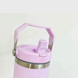 Puede incluir: Una taza de viaje de acero inoxidable de color lavanda con tapa y asa de transporte. La tapa tiene un pico abatible.