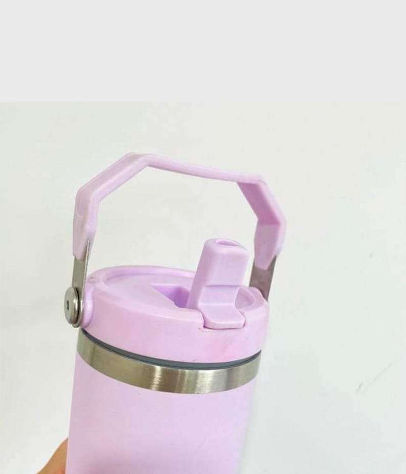 Puede incluir: Una taza de viaje de acero inoxidable de color lavanda con tapa y asa de transporte. La tapa tiene un pico abatible.