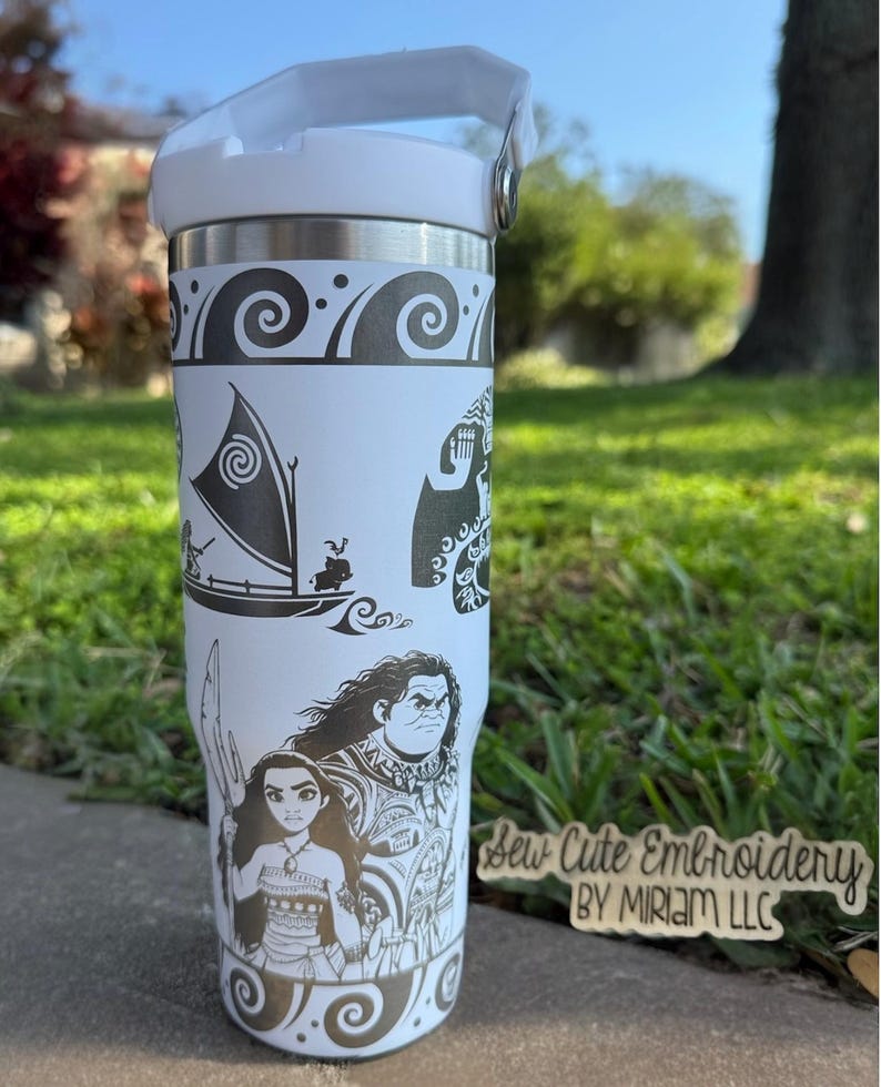 May include: White stainless steel tumbler with a black and white design featuring characters from the animated film Moana. The design includes Moana, Maui, and a sailing canoe. The tumbler has a black lid with a carrying handle. The text "Sew Cute Embroidery by Miriam LLC" is visible in the lower right corner of the image.