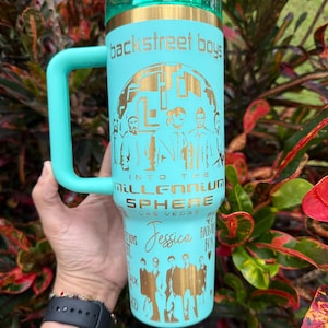 May include: A teal insulated travel mug with a handle, featuring gold text and graphics. The text includes "Backstreet Boys," "Into the Millennium Sphere," and "Jessica." The mug has a teal lid and handle.