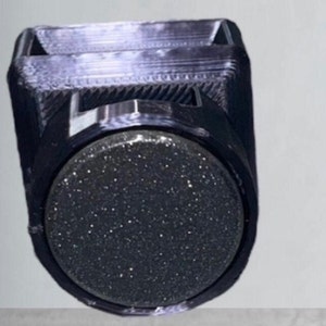 May include: A black 3D printed holder featuring a square top opening and a circular front face. The circular front panel has a dark, glittery, textured surface that contrasts with the smooth, layered finish of the surrounding black plastic frame.