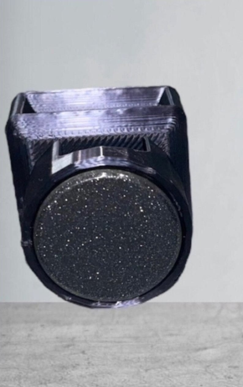 May include: A black 3D printed holder featuring a square top opening and a circular front face. The circular front panel has a dark, glittery, textured surface that contrasts with the smooth, layered finish of the surrounding black plastic frame.