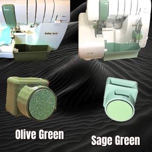 May include: Two Baby Lock serger machines featuring custom attachments in olive green and sage green. The olive green attachment has a textured, glittery circular face, while the sage green attachment has a smooth, matte circular face.