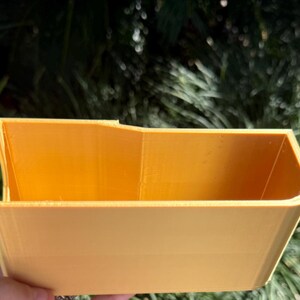 Op de afbeelding: Een kleine, rechthoekige container met een open bovenkant in een warme gele kleur. De container heeft een licht trapeziumvormige vorm, met schuine zijkanten en een platte bodem. Het object wordt tegen een achtergrond van groen gebladerte gehouden.