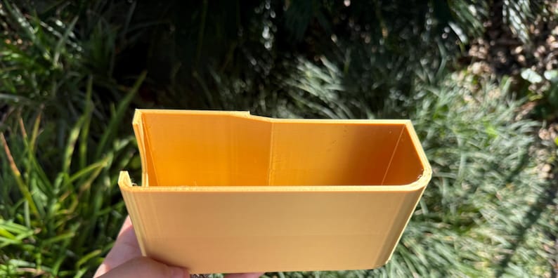 Op de afbeelding: Een kleine, rechthoekige container met een open bovenkant in een warme gele kleur. De container heeft een licht trapeziumvormige vorm, met schuine zijkanten en een platte bodem. Het object wordt tegen een achtergrond van groen gebladerte gehouden.
