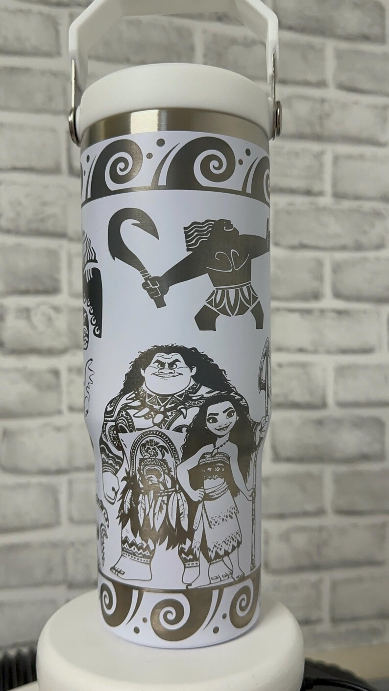 May include: White stainless steel travel tumbler with a black and white design featuring characters from the animated movie Moana. The design includes Maui, Moana, and a hook.