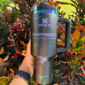 May include: Black Stanley travel mug with a handle, featuring the Embry-Riddle Aeronautical University logo and airplane graphics. The mug has a black lid with iridescent accents. The text "STANLEY" is visible.