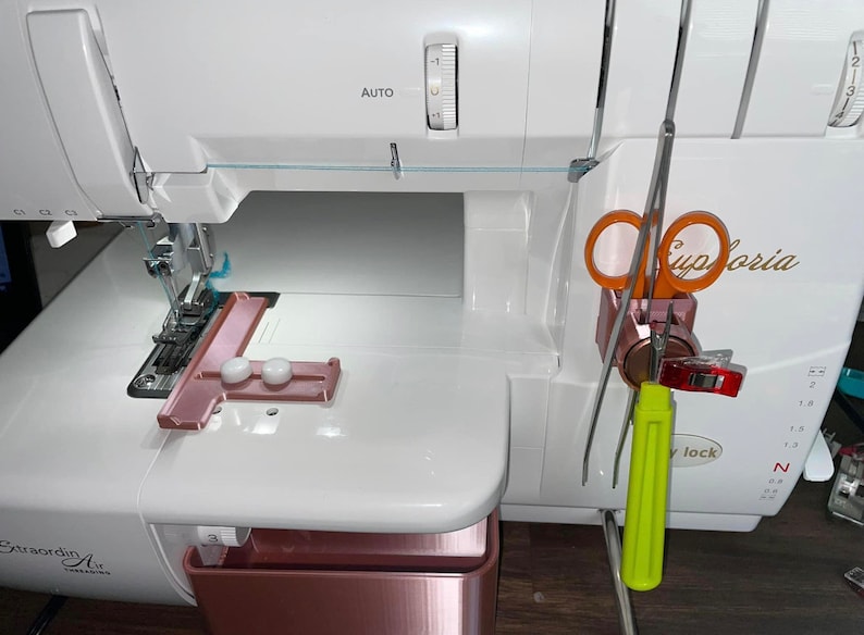 May include: A white sewing machine featuring a rose gold accessory tray and a matching rose gold tool holder attached to the side. The holder contains orange scissors, a lime green seam ripper, and a red plastic sewing clip for organization.