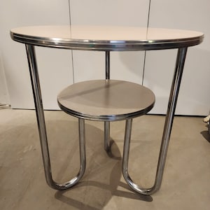 Könnte beinhalten: Ein runder Beistelltisch aus Chrom und Weiß mit einem unteren Regal. Der Tisch hat ein schlichtes, modernes Design mit geschwungenen Beinen.