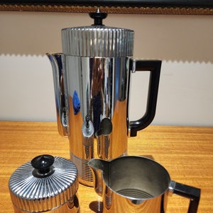 Könnte beinhalten: Ein silbernes Kaffeekännchen, Milchkännchen und Zuckerdose mit schwarzen Griffen. Das Set hat ein geripptes Design und eine polierte Oberfläche.