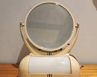May include: Vintage cream-colored vanity mirror with a round, pivoting mirror and a rectangular light fixture. The base features vertical brown stripes. The mirror is set on a wooden surface.