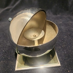 May include: A silver, round, two-piece ashtray with a small, round, silver insert in the center. The ashtray is sitting on a green and silver square base.
