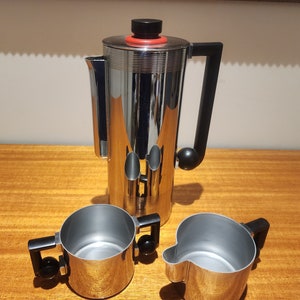 Könnte beinhalten: Ein Set aus drei silberfarbenen Kaffee- oder Teeservice-Teilen. Das Hauptstück ist ein hoher, zylindrischer Krug mit einem schwarzen Knopf auf dem Deckel. Die beiden anderen Teile sind kleinere, runde Tassen mit schwarzen Griffen.