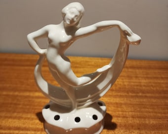May include: White ceramic figurine of a woman dancing, holding a flowing piece of fabric. The figurine is on a base with holes for flower stems, making it a decorative flower vase.