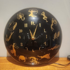 May include: A black and gold clock with Roman numerals and zodiac symbols around the face. The clock is round and has a dome-shaped glass cover.