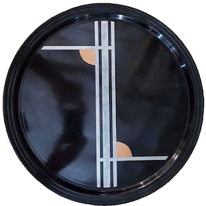 Puede incluir: Una bandeja circular negra con un diseño geométrico plateado y dorado. La bandeja tiene un borde elevado.