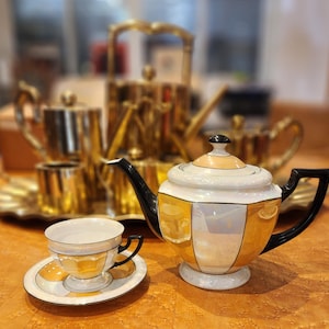 Puede incluir: Una tetera dorada y blanca con detalles negros y una taza y un platillo a juego. La tetera tiene un diseño geométrico con un acabado perlado.