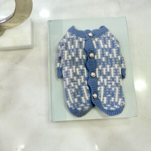 May include: A blue and white patterned dog sweater with pearl-like buttons. The sweater is made of a soft knit material and is designed to fit a small dog. The sweater is lying on a light blue book.