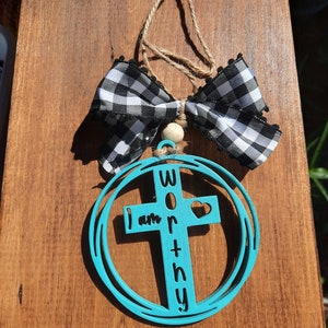 May include: A teal blue wooden ornament shaped like a circle with a cross inside. The cross has the words "I am worthy" written on it. The ornament is decorated with a black and white checkered bow.