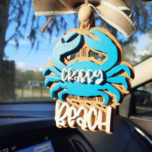 May include: Wooden crab-shaped car air freshener with blue accents and the words "Crabby Please Return To Beach". It is suspended by a beige ribbon and twine, hanging in front of a car dashboard.
