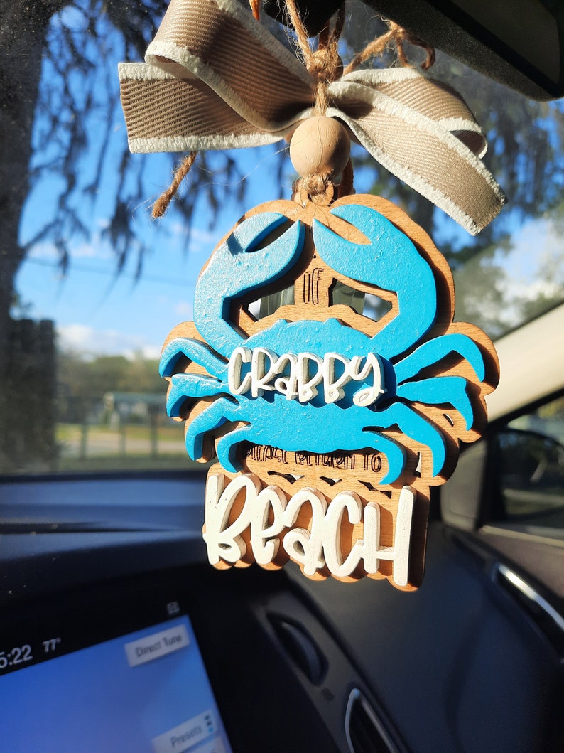 May include: Wooden crab-shaped car air freshener with blue accents and the words "Crabby Please Return To Beach". It is suspended by a beige ribbon and twine, hanging in front of a car dashboard.