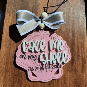 May include: A pink seashell-shaped ornament with the text "Call me on my shell, I'll be at the beach" in white script. It has a beige and white ribbon bow and wooden beads. A black string is attached for hanging.