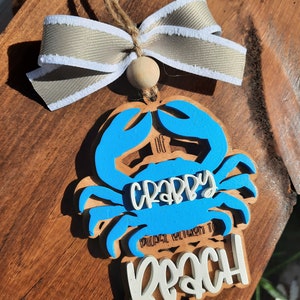 May include: Wooden crab ornament with blue accents and the words "Crappy" and "Beach" in white. The ornament has a beige ribbon and wooden bead for hanging. A fun beach-themed decoration.