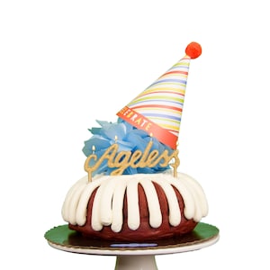 Puede incluir: Un pastel bundt de terciopelo rojo con glaseado blanco y una decoración de purpurina dorada "Ageless". El pastel está decorado con un pompón de papel de seda azul y un gorro de fiesta a rayas de colores con la palabra "Celebrate" en él.