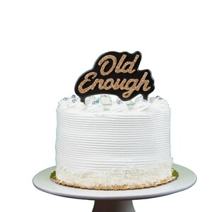 Puede incluir: Pastel blanco con un adorno negro y dorado con purpurina que dice "Old Enough".