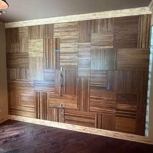 May include: A wall covered in rectangular wood panels featuring a vertical and horizontal striped pattern. The panels are arranged in a grid-like mosaic design with varying orientations. Small blue painter's tape markers are visible on the wall.