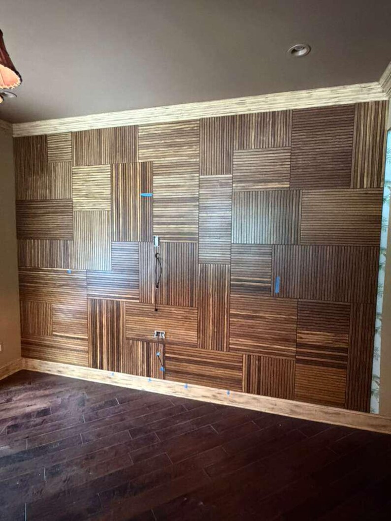May include: A wall covered in rectangular wood panels featuring a vertical and horizontal striped pattern. The panels are arranged in a grid-like mosaic design with varying orientations. Small blue painter's tape markers are visible on the wall.
