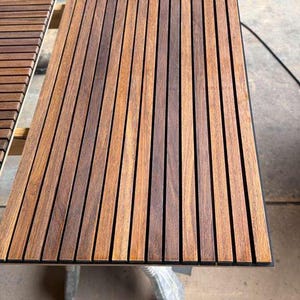 May include: A rectangular acoustic wall panel featuring vertical wooden slats in a medium brown tone with visible wood grain, separated by narrow black felt gaps. The panel has a clean, linear design with uniform spacing between each wooden strip.