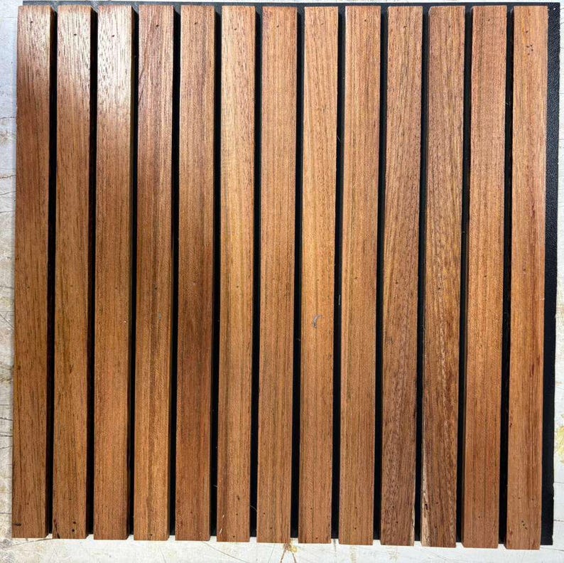 Op de afbeelding: Een vierkant houten paneel met verticale, gelijkmatig verdeelde, roodbruine houten latten. De lamellen worden gescheiden door dunne, zwarte lijnen, waardoor een gestreept patroon ontstaat. De houtnerf is zichtbaar en voegt textuur toe aan het oppervlak.