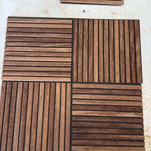 Op de afbeelding: Vier vierkante houten terrasplanken met een lattenontwerp, gerangschikt op een lichtgekleurd oppervlak. De tegels zijn gemaakt van donkerbruin hout met zwarte randen. Extra houten latten zijn in de buurt opgestapeld.