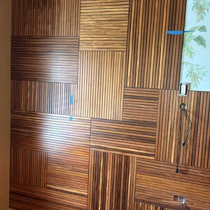 May include: A wall installation featuring rectangular panels of dark brown wood slats arranged in a geometric grid pattern. The slats are oriented both horizontally and vertically, partially covering a light blue wallpaper with a green palm leaf print.