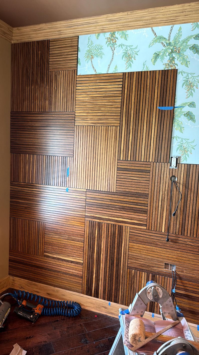 May include: A wall installation featuring rectangular panels of dark brown wood slats arranged in a geometric grid pattern. The slats are oriented both horizontally and vertically, partially covering a light blue wallpaper with a green palm leaf print.