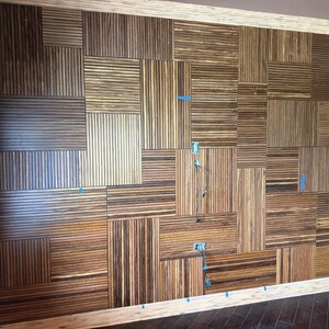 May include: A wall installation composed of rectangular wooden panels featuring a striped pattern of dark brown and light tan wood grain. The panels are arranged in a grid with varying orientations, and small blue tape markers are visible.